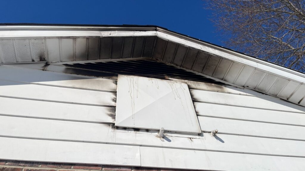 The corner of a house shows blackened marks and streaks below a blocked vent, indicative of recent fire damage against a clear blue sky.