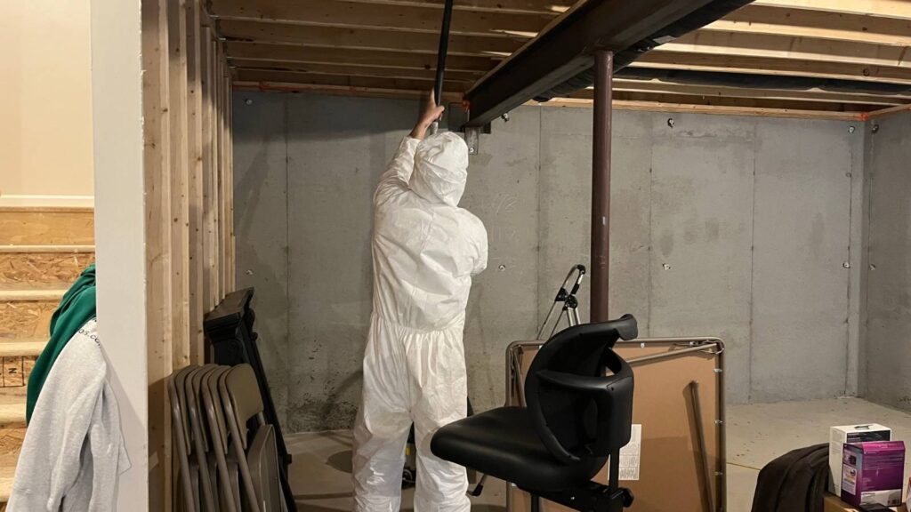 A person in a white protective suit reaches up to adjust a pipe in a dimly lit basement filled with furniture and construction materials.