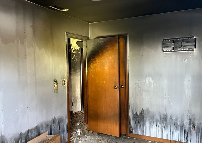 A room with charred walls and a blackened door, indicating significant fire damage, with soot stains and debris on the floor.
