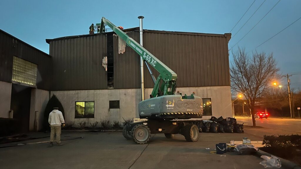 A green aerial lift elevates workers next to a building at dusk, with construction debris and bags in the foreground.