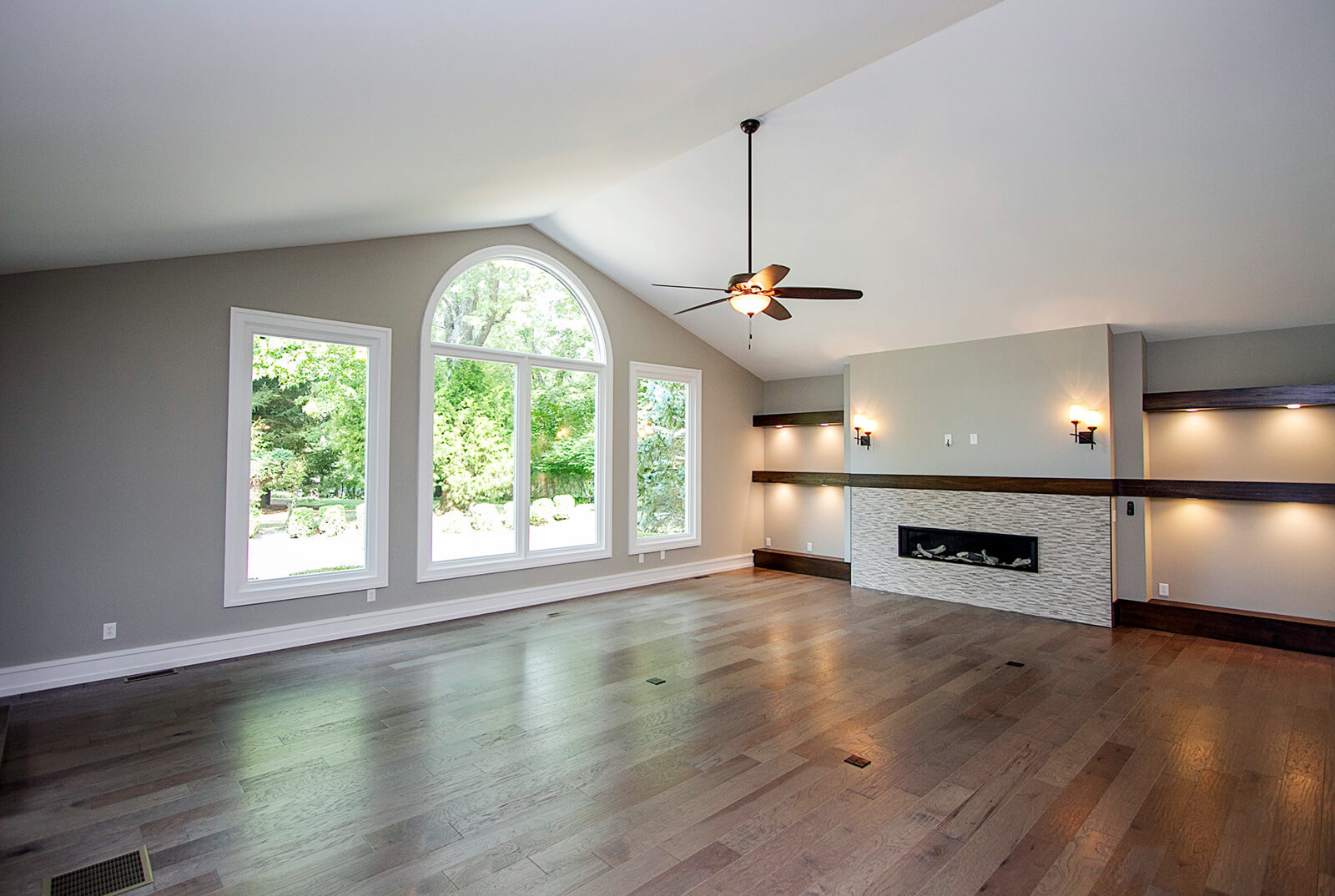 Modern living room with vaulted ceiling, large arched window, and rectangular windows overlooking green trees. The room features a light gray wall, wooden floor, a fireplace with a textured white tile surround, and dark wood mantels with recessed lighting.